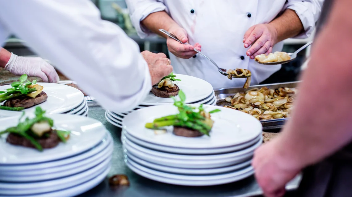 Chefs plating dishes on a stainless steel pass in a commercial kitchen