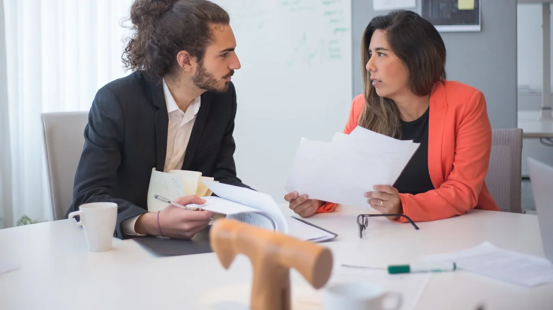 Two colleagues comparing paperwork across a table in a meeting room
