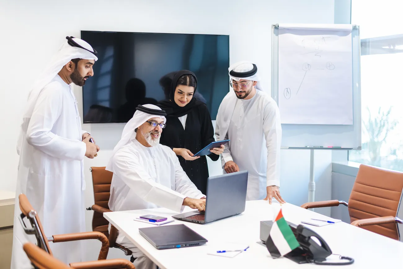 A team of Emirati professionals reviewing company filings around a laptop in a Dubai office